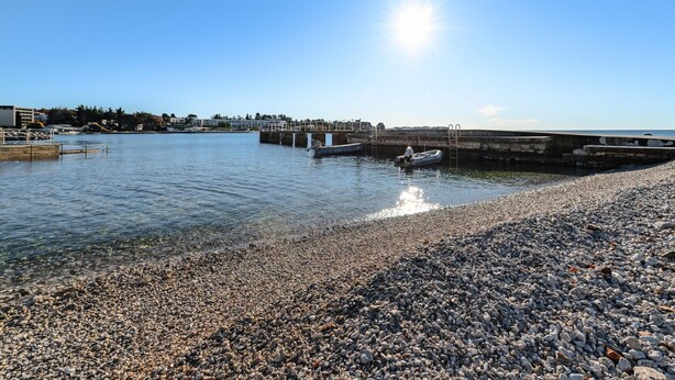 A pebbled beach with a boat docked near a pier, clear skies, and buildings in the background. - Pretty Apartment With Balcony (Porec)