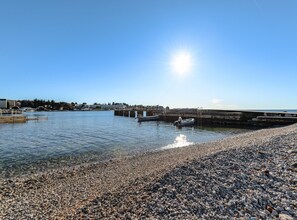 A pebbled beach with a boat docked near a pier, clear skies, and buildings in the background. - Pretty Apartment With Balcony (Porec)