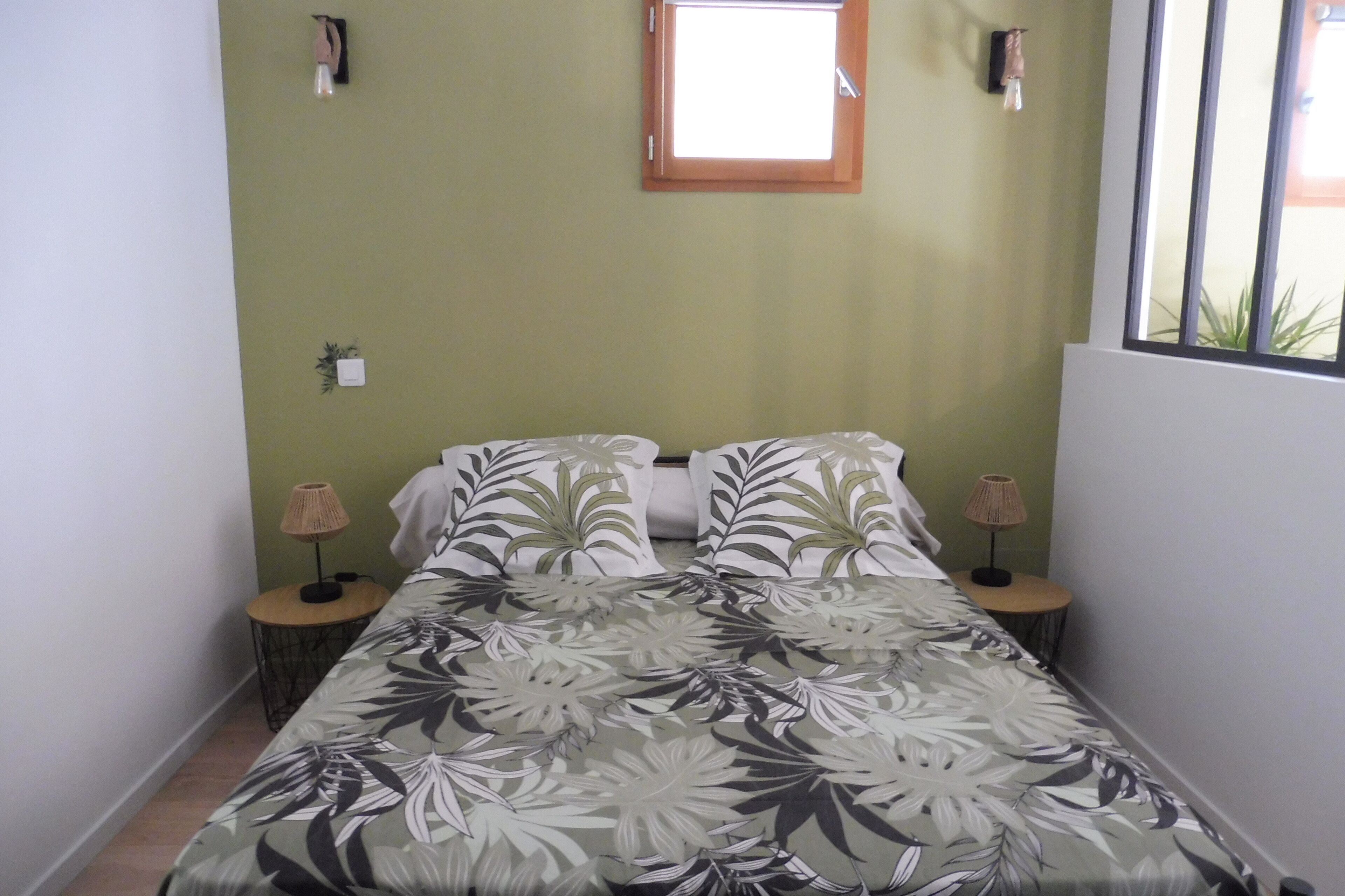 Une chambre à coucher avec une couvre-lit à motifs floraux, deux tables de chevet avec des lampes, une fenêtre et une petite plante accrochée au mur.
