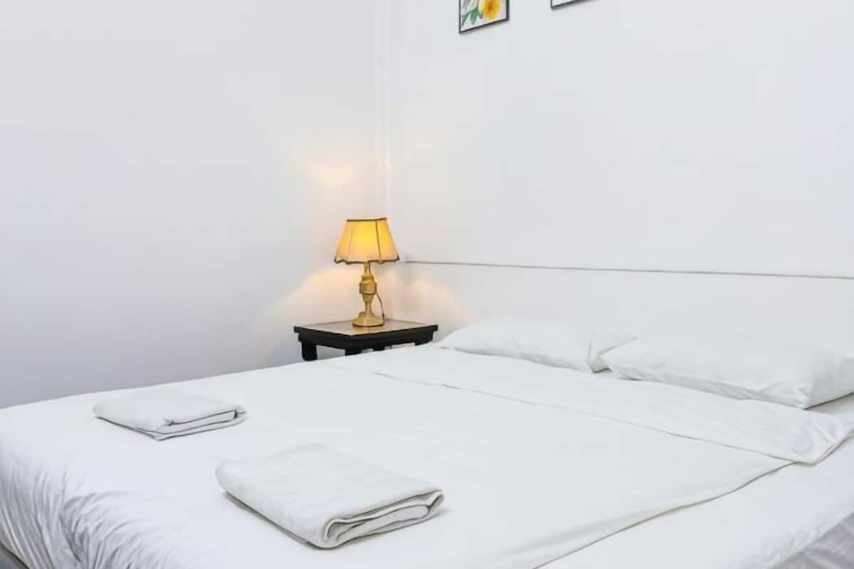 Una cama bien tendida con sábanas blancas y una toalla doblada, una lámpara de noche y una obra de arte colgada en la pared.