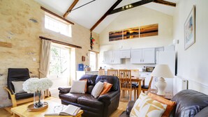 A cozy living room with a leather sofa, wooden coffee table, and a dining area with wooden chairs. A kitchen with white cabinets is visible in the background.