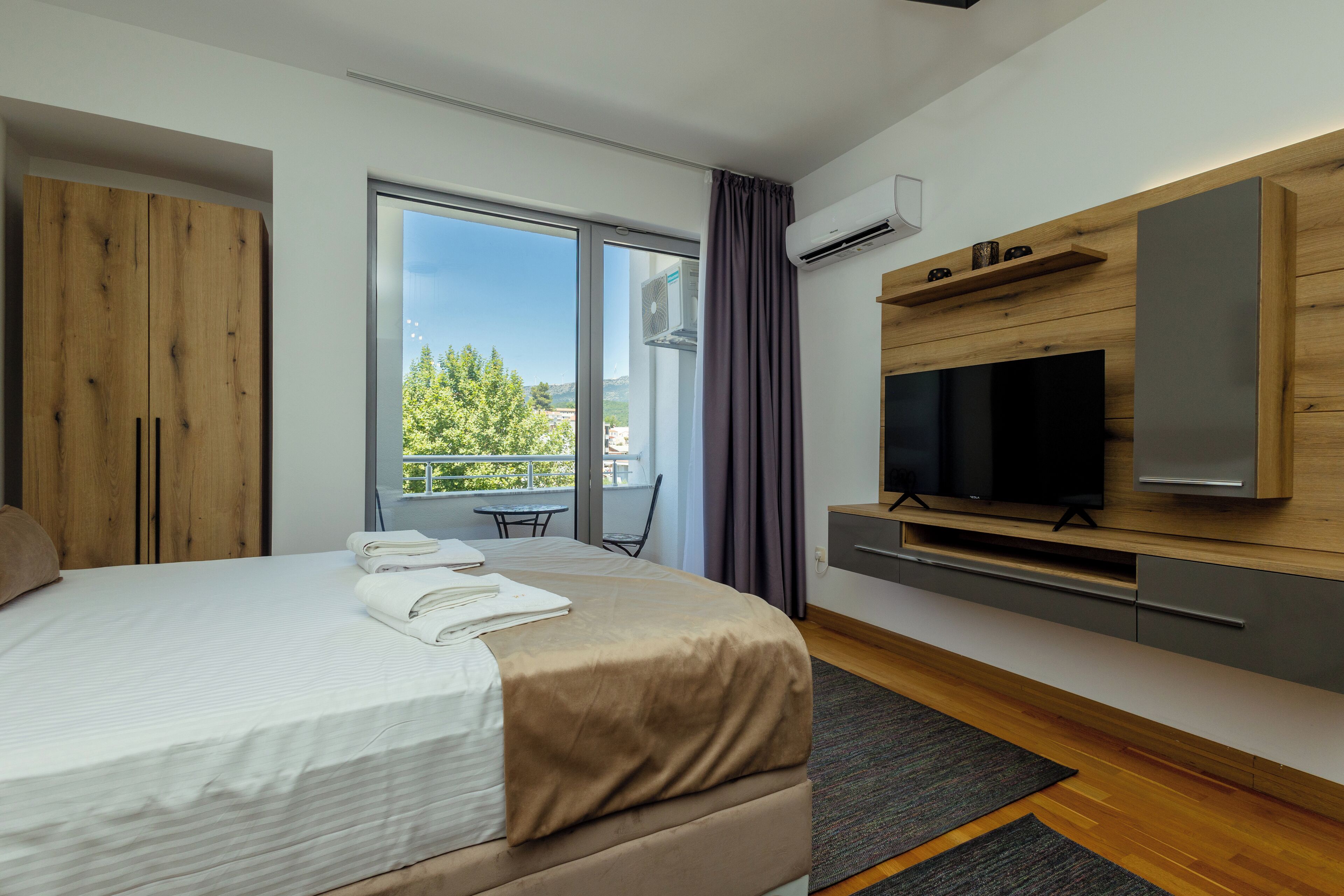 Ein modernes Hotelzimmer mit einem großen Bett, einem Fernseher, einem Holzschrank und einem Balkon mit Blick auf Bäume und Gebäude.