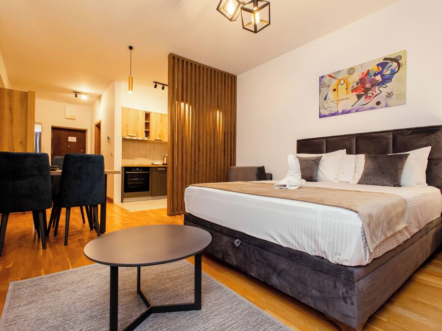 Una habitación de hotel moderna con una cama grande, una zona de comedor con sillas, una mesita, una cocina y un cuadro en la pared.