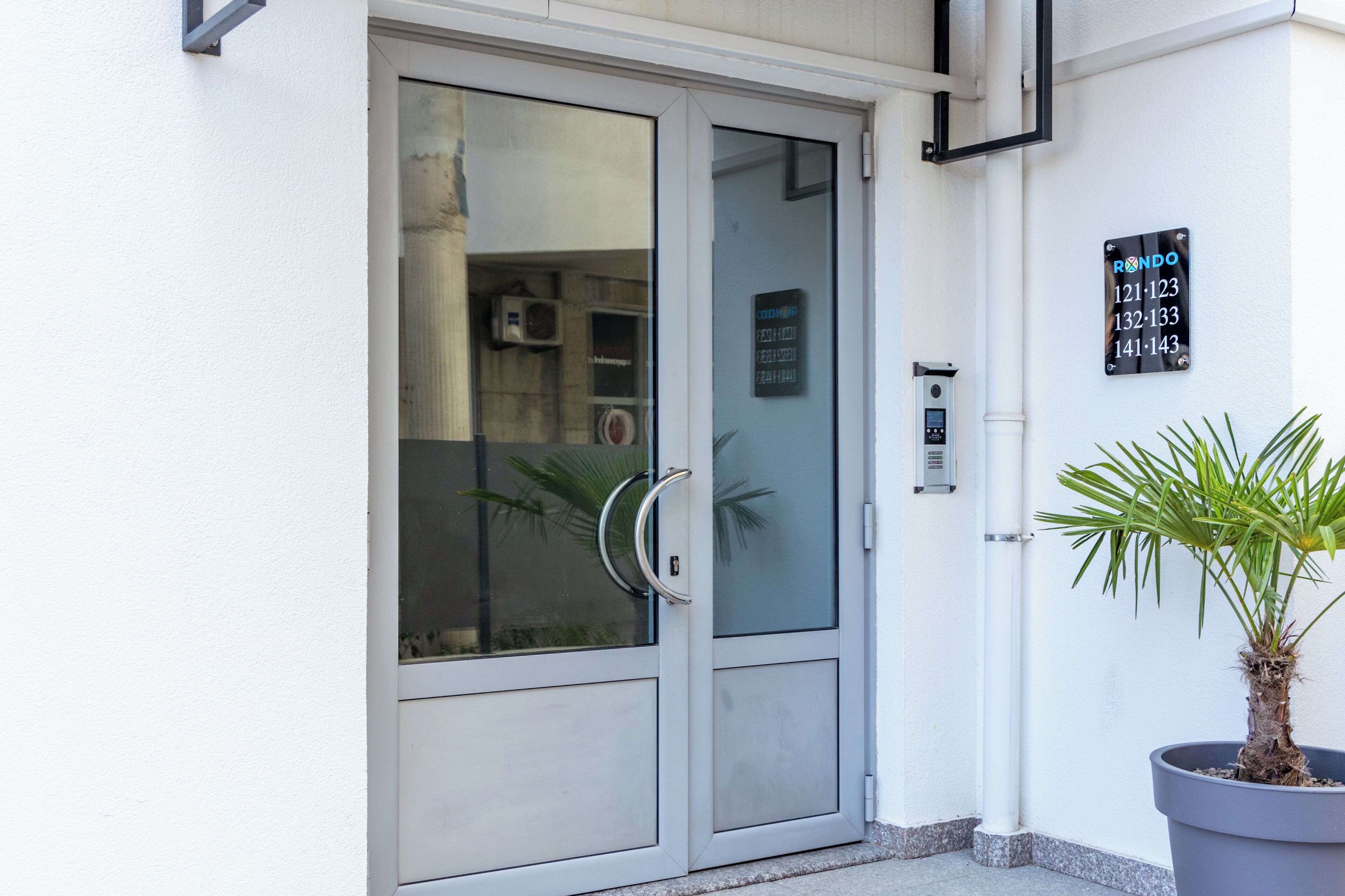 A modern building entrance with a glass door, a potted plant, and a sign listing apartment numbers.