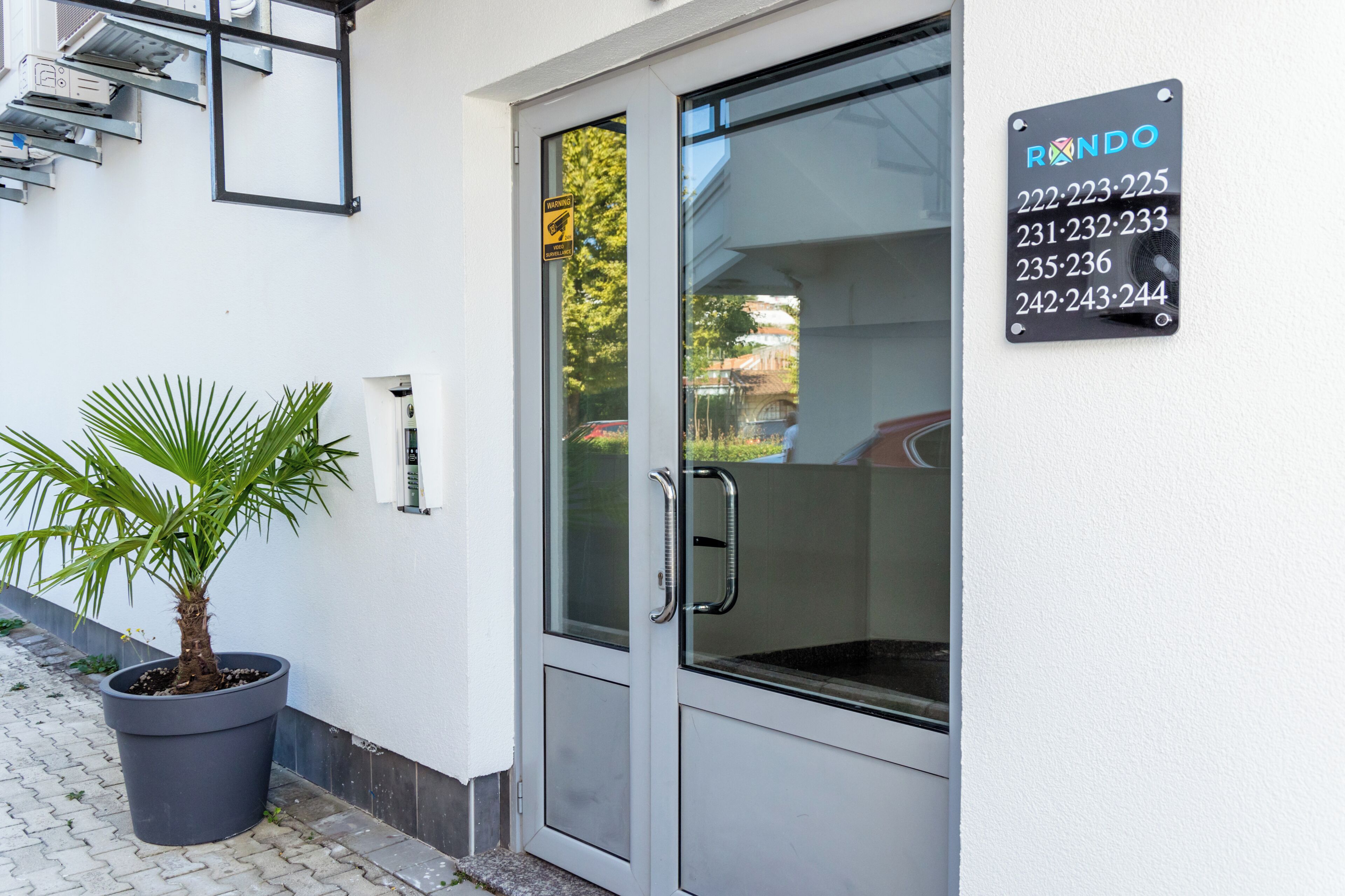 Una entrada moderna de edificio con puerta de vidrio, planta en maceta y cartel con números de teléfono.