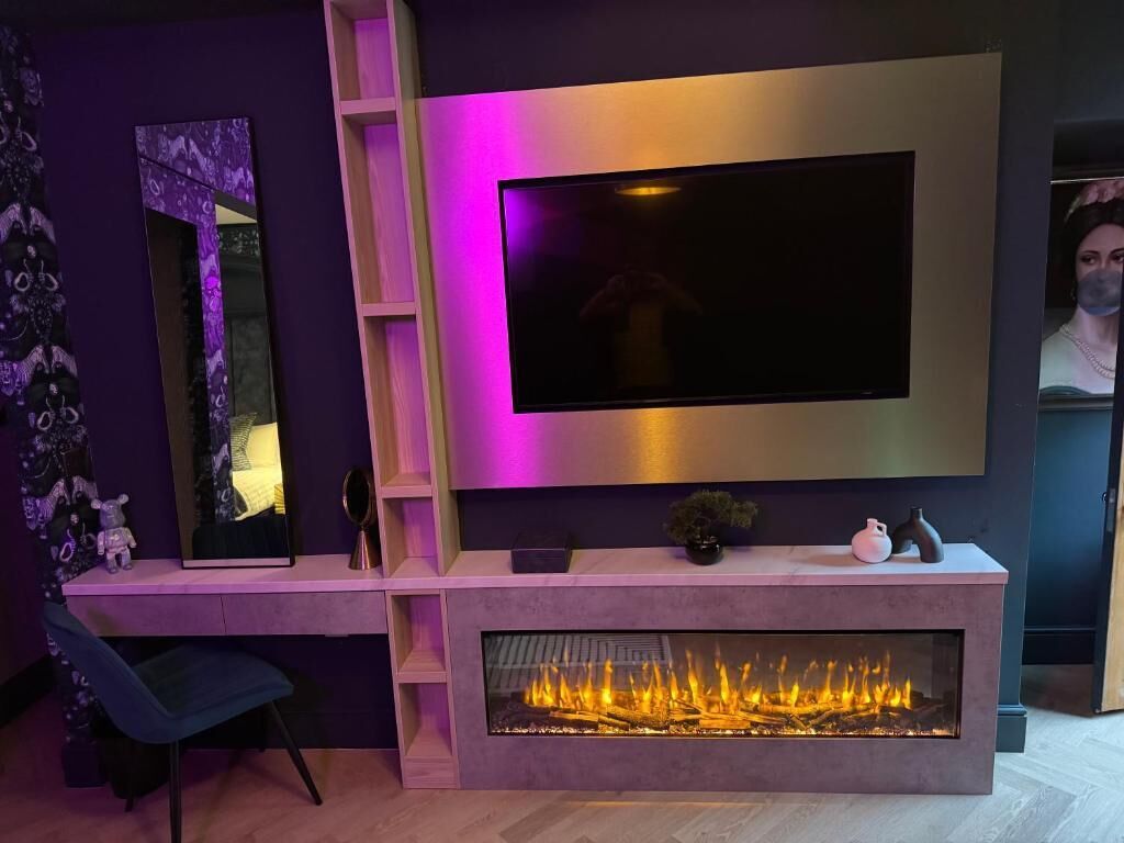 A modern living room with a flat-screen TV mounted on the wall, a built-in fireplace, a wooden entertainment unit, and a chair.