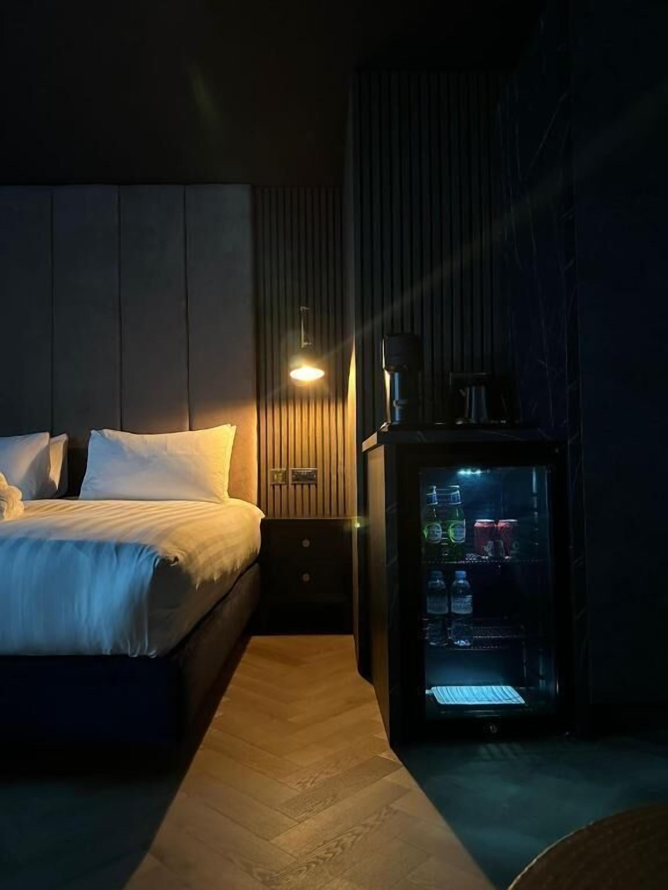 Habitación de hotel con cama, mesita de noche, lámpara y minibar.