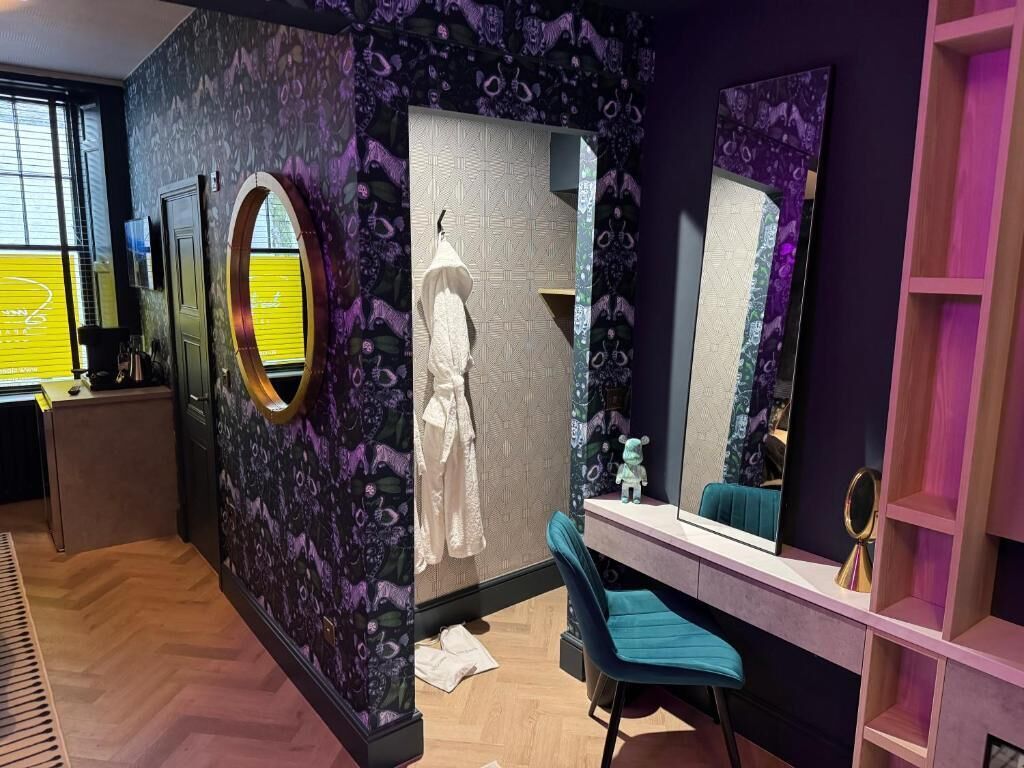 A room with a mirror, a chair, a vanity with a mirror, and a wardrobe.