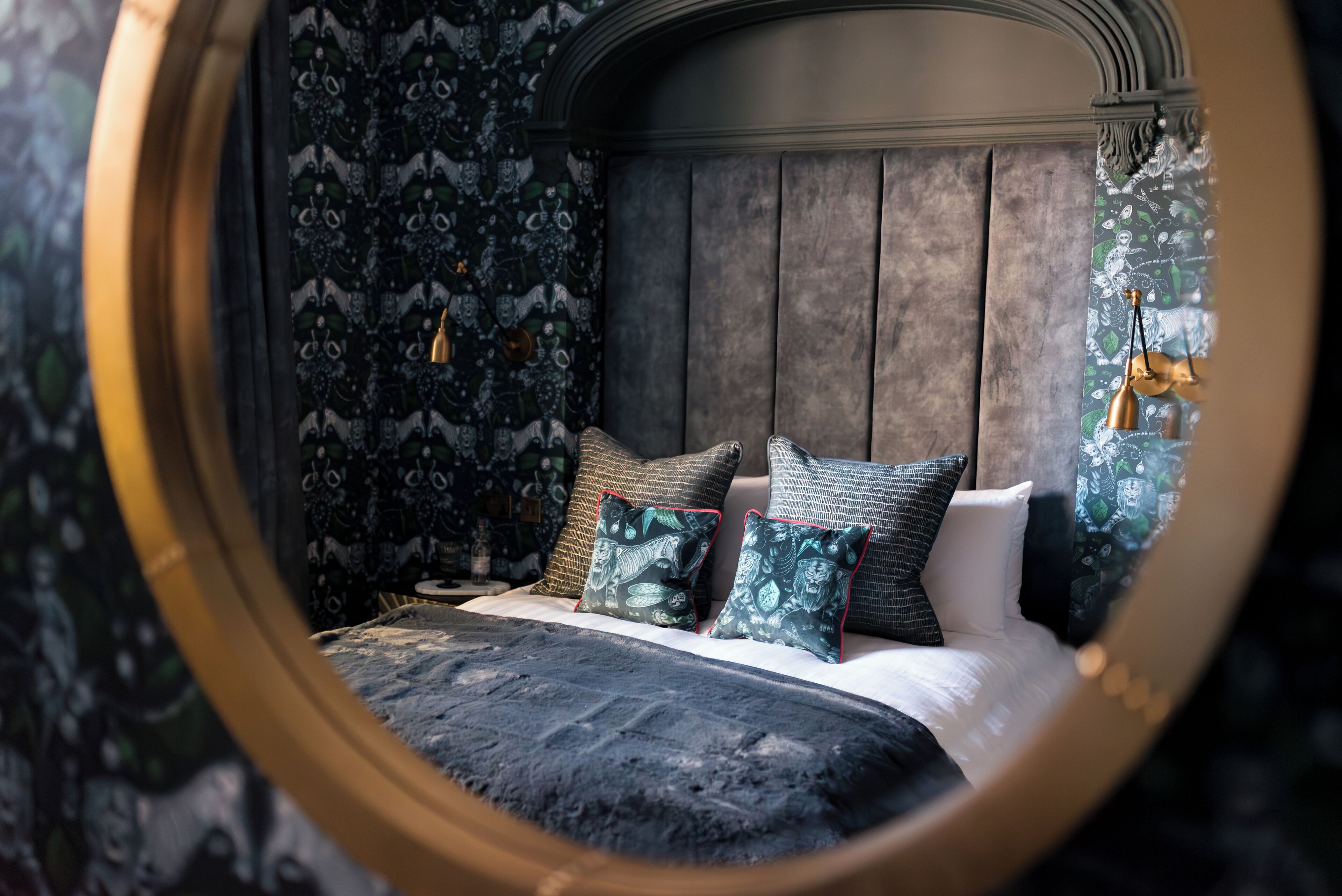 A bedroom with a round mirror reflecting the bed and patterned wallpaper.