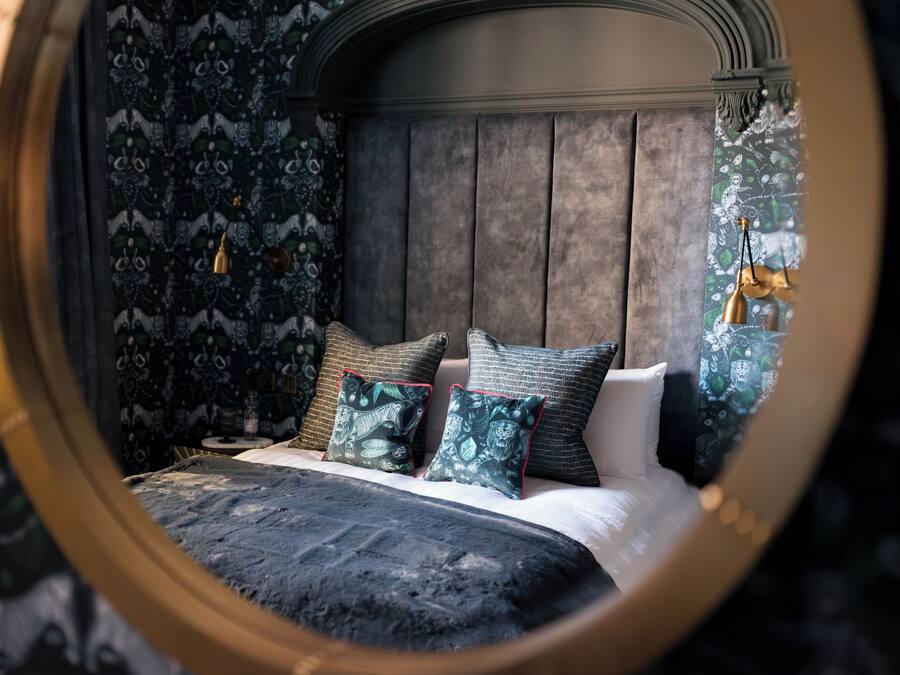 A bedroom with a round mirror reflecting the bed and patterned wallpaper.