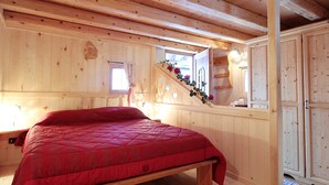 A wooden cabin bedroom with a bed, a window with curtains, a staircase with a floral decoration, and a wardrobe.