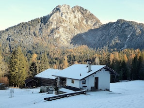 A house with a snow-covered roof, a chimney, and a wooden deck in a snowy landscape with a forest and mountains in the background. - Baita Mountain View Pale San Martino (Primiero San Martino di Castrozza)