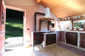 A kitchen with a stone-tiled floor, wooden cabinets, a stove, and a view of a garden through the open door. - Baita Mountain View Pale San Martino (Primiero San Martino di Castrozza)