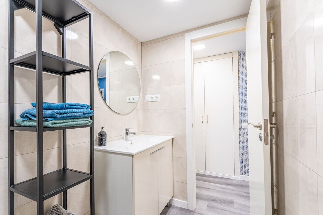 Une salle de bain avec un lavabo blanc, un miroir, un porte-serviettes avec des serviettes pliées et une porte donnant sur une autre pièce.