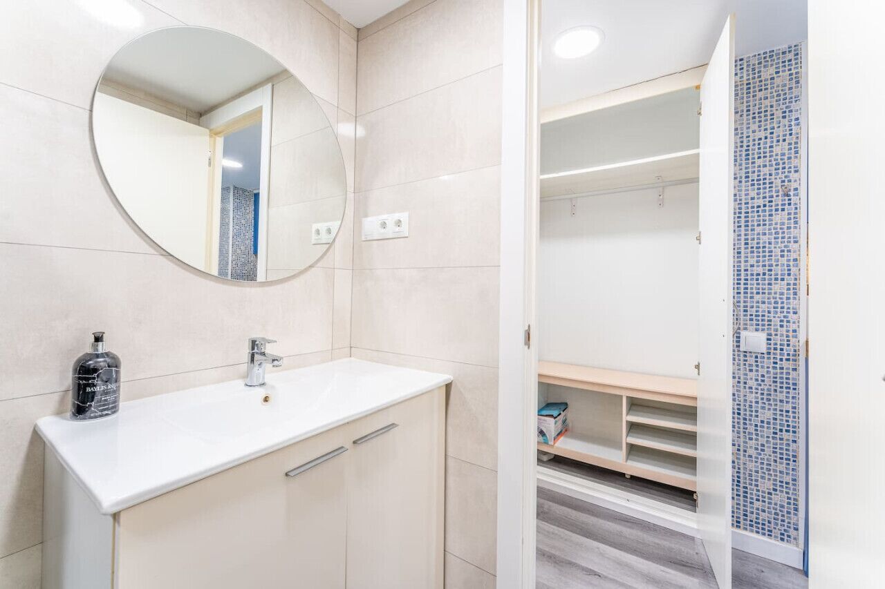 Un baño con un espejo redondo, un lavabo blanco y una botella de jabón líquido.
