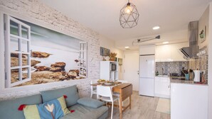 A modern kitchen with white cabinets, a dining area with a wooden table and chairs, and a living room with a sofa and a wall-mounted TV. - Salou Luxe Stay | Chill-out | Cozy ( 2BR ) Parking (Salou)