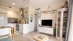 A modern kitchen with white cabinets, a dining area with a table and chairs, a living room with a TV and a white cabinet, and a small round table with magazines. - Salou Luxe Stay | Chill-out | Cozy ( 2BR ) Parking (Salou)