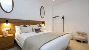 A modern bedroom with a wooden headboard, a large bed with multiple pillows, a round mirror, a black metal rack, and a small round stool.