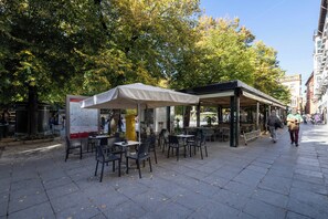 Un café al aire libre con mesas y sillas, rodeado de árboles y edificios.
