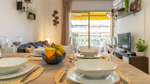 Un coin repas avec une table dressée pour quatre personnes, un saladier de fruits et un téléviseur en arrière-plan.