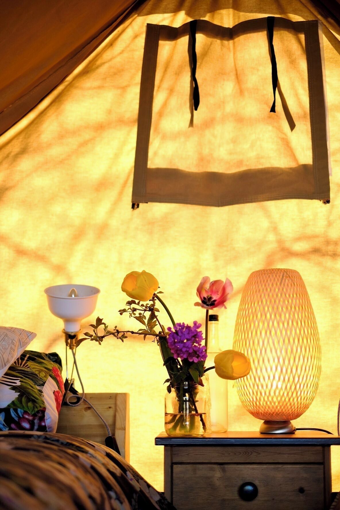 Interior de una tienda de campaña con una cama, una mesita de noche con lámpara, un florero con flores y una lámpara colgante.