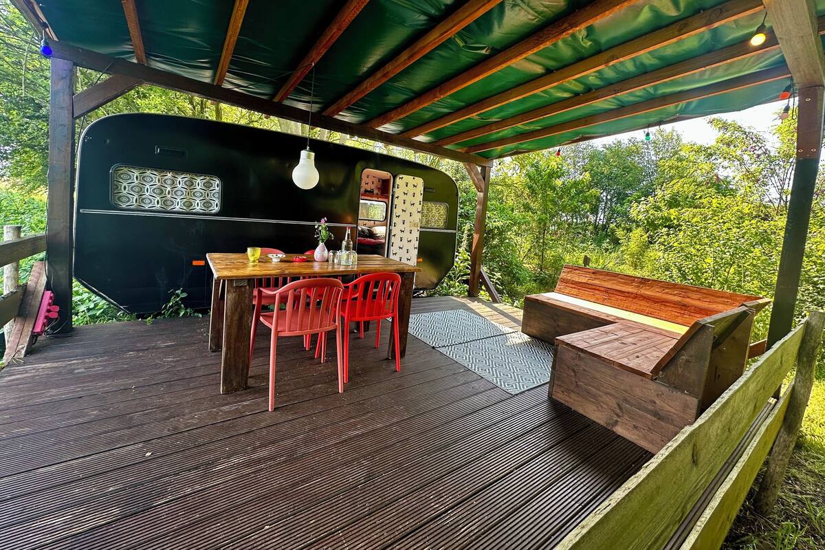 A covered outdoor seating area with a table and chairs, a bench, and a camper van in the background.