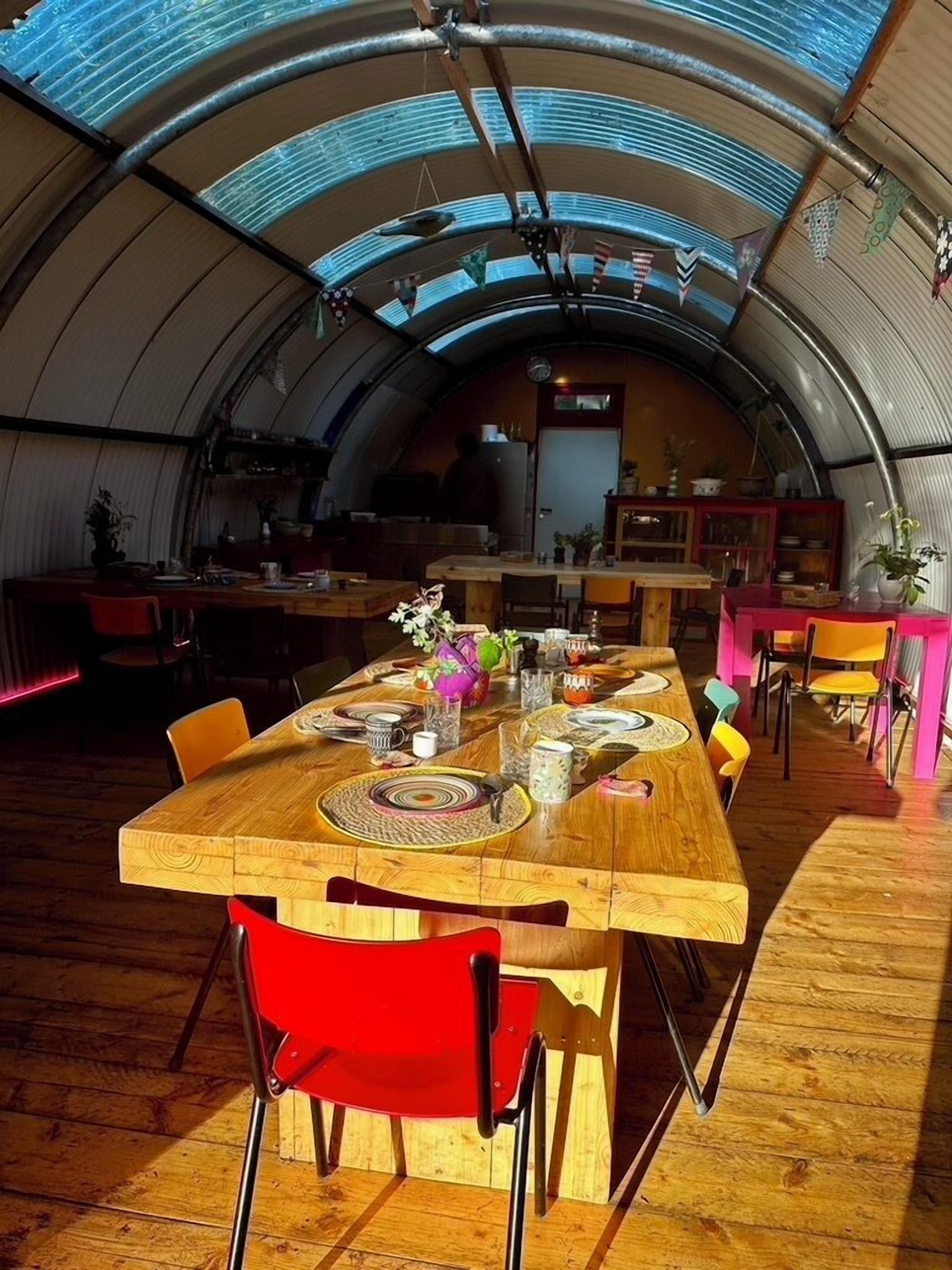 A spacious, curved-roofed interior with a long wooden dining table set for a meal, surrounded by colorful chairs.