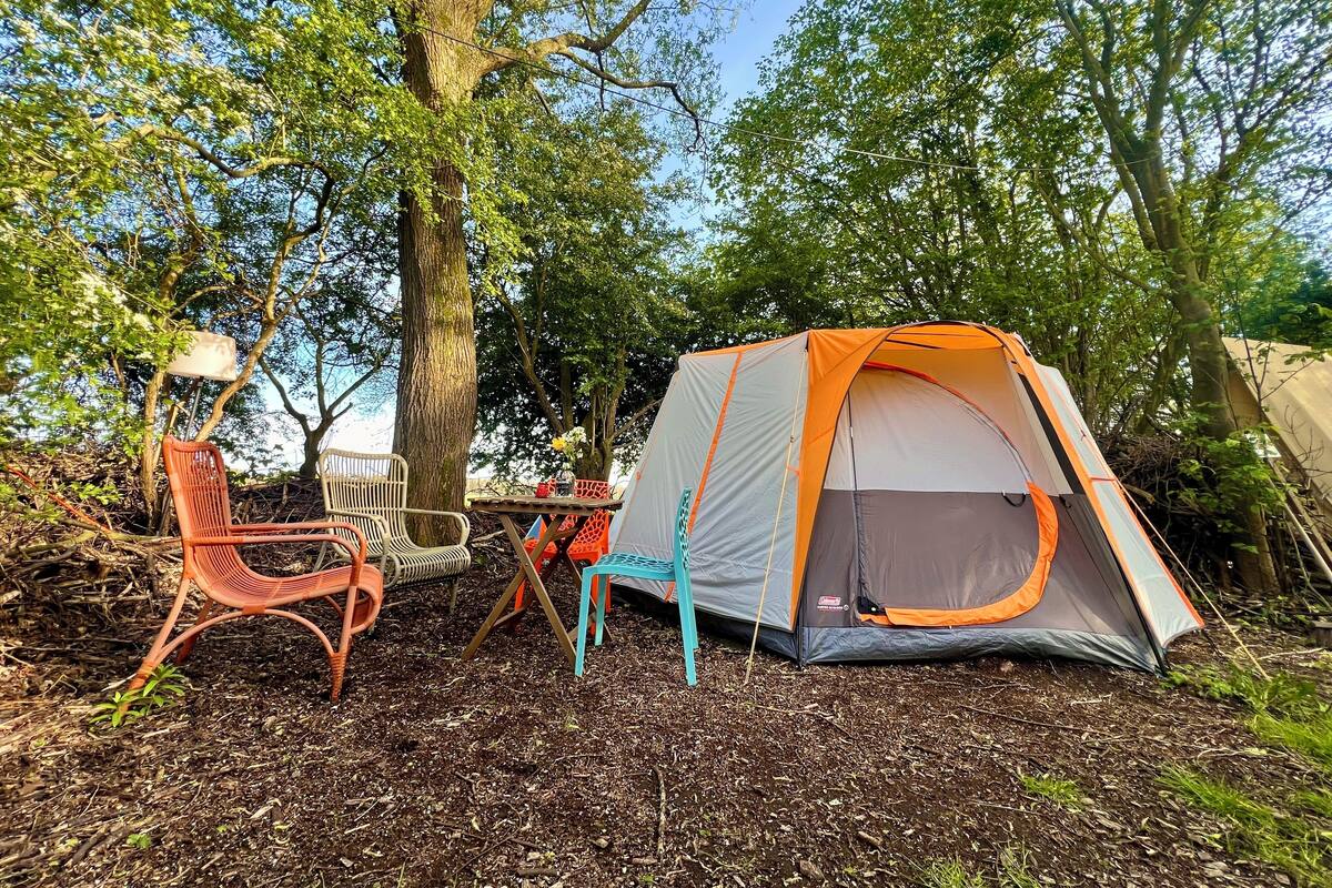 A tent set up in a wooded area with a table and chairs nearby.