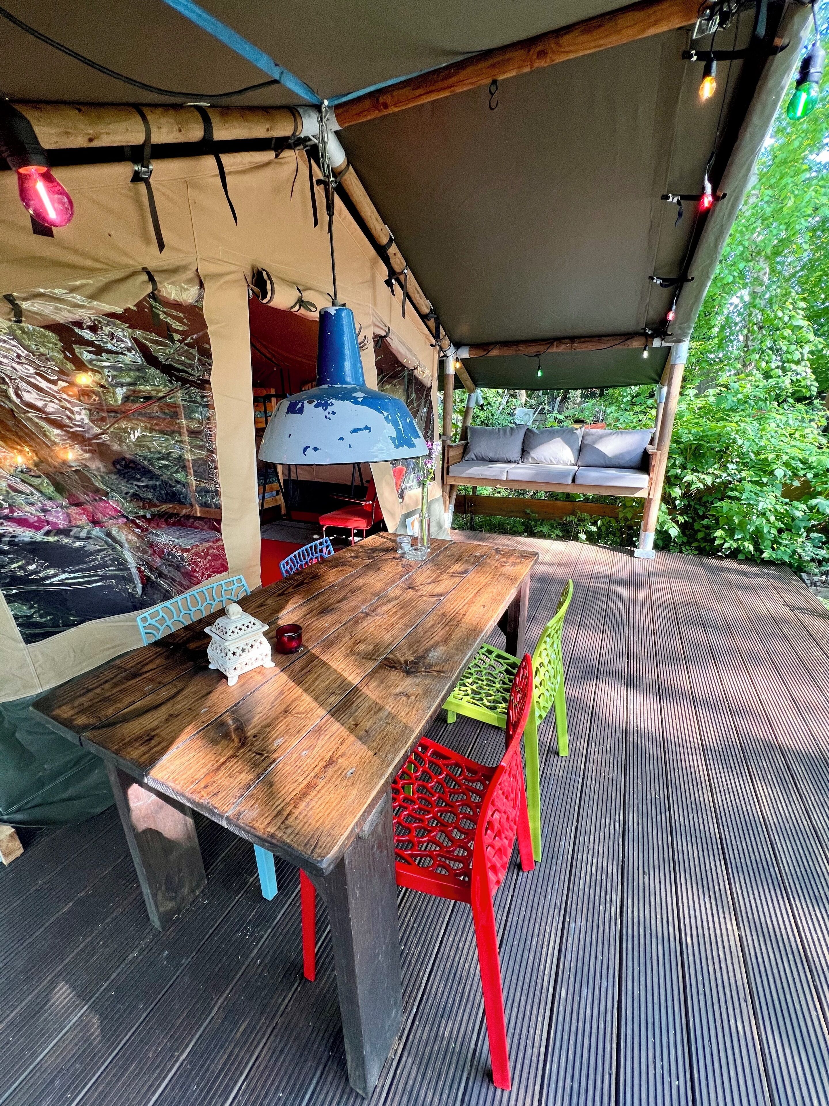 Eine Holzterrasse mit Tisch und Stühlen, einer hängenden Leuchte und einem überdachten Bereich mit einer gepolsterten Bank.