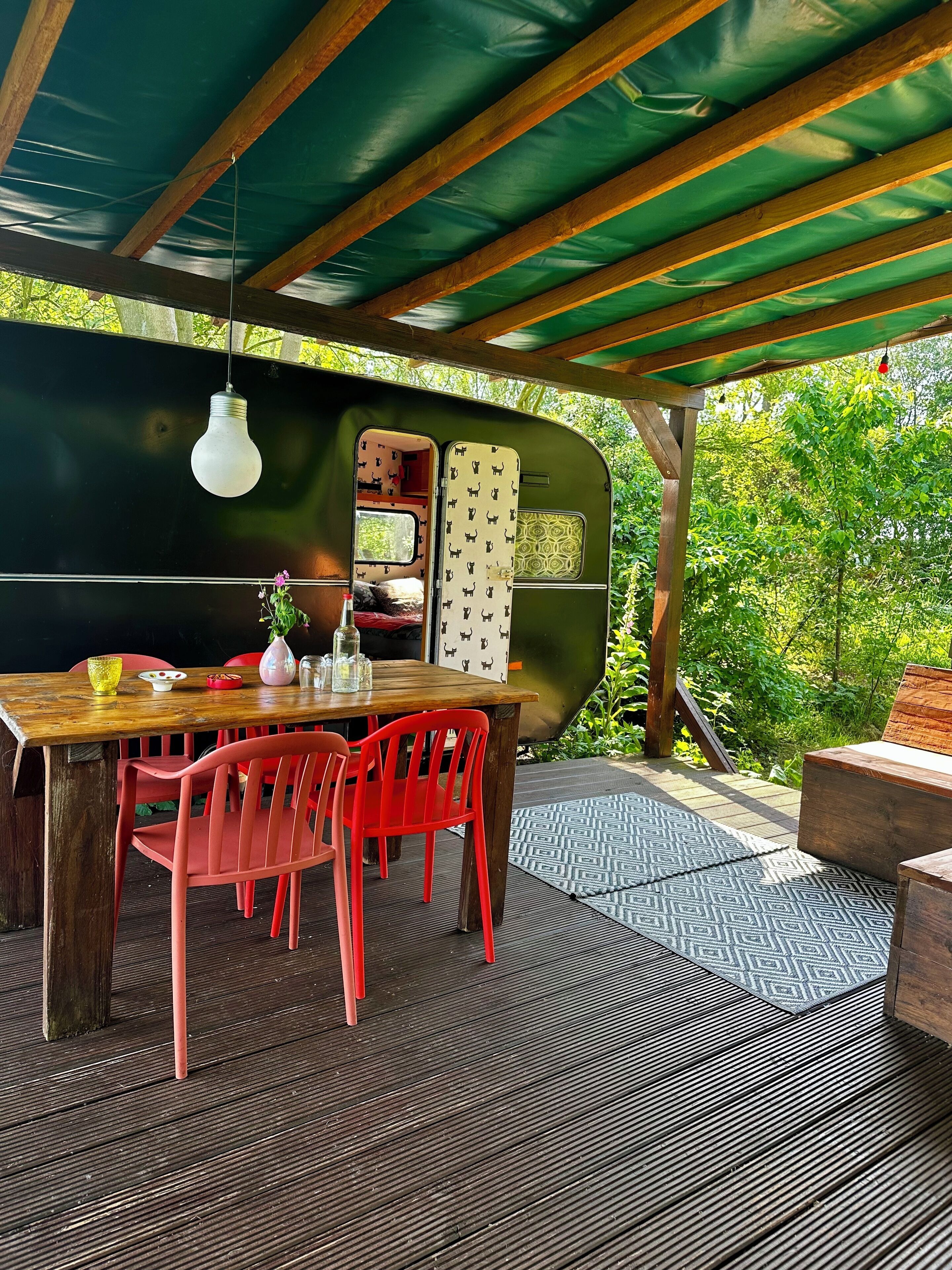 A covered outdoor dining area with a wooden table and red chairs, a black grill, and a green awning.
