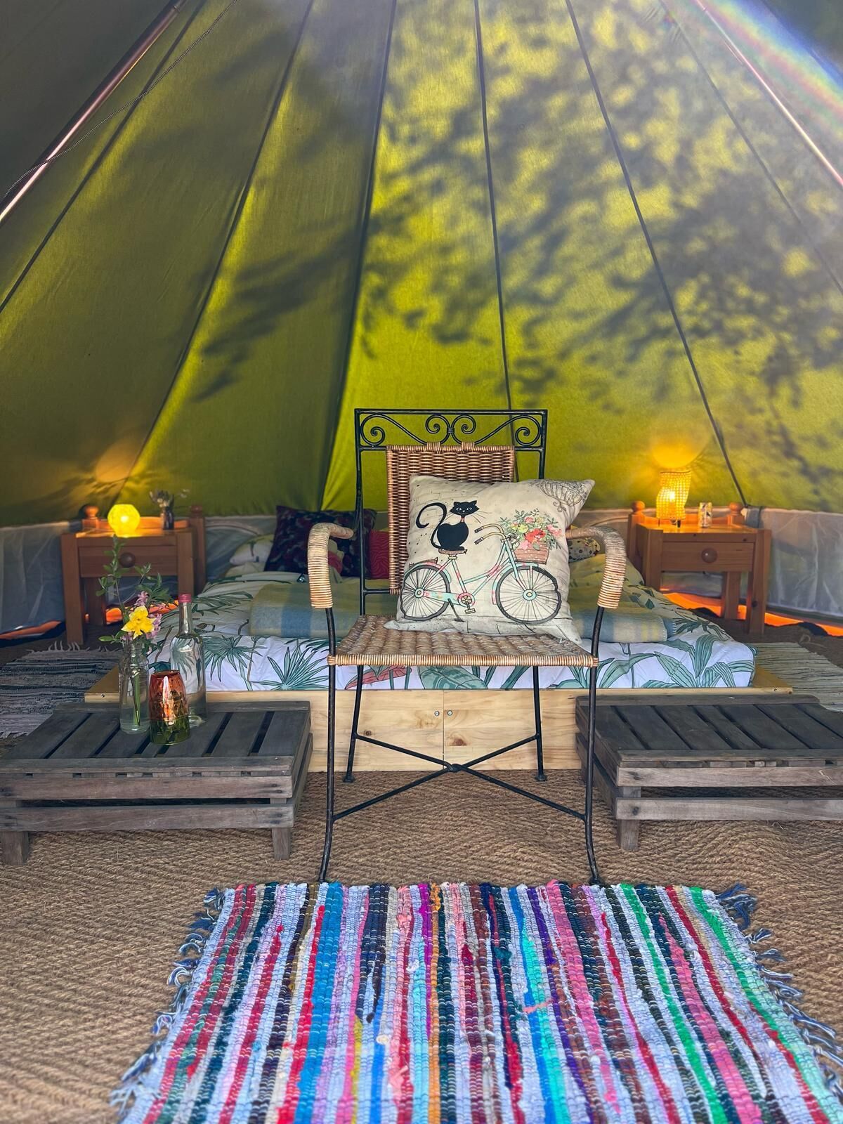 A cozy tent interior with a bed, a chair, a colorful rug, and two nightstands with lamps.