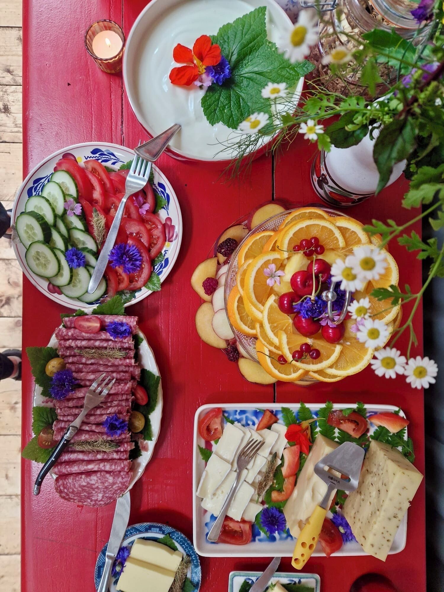 Una mesa con diversos alimentos, como pepinos en rodajas, tomates, naranjas y queso, sobre un mantel rojo.