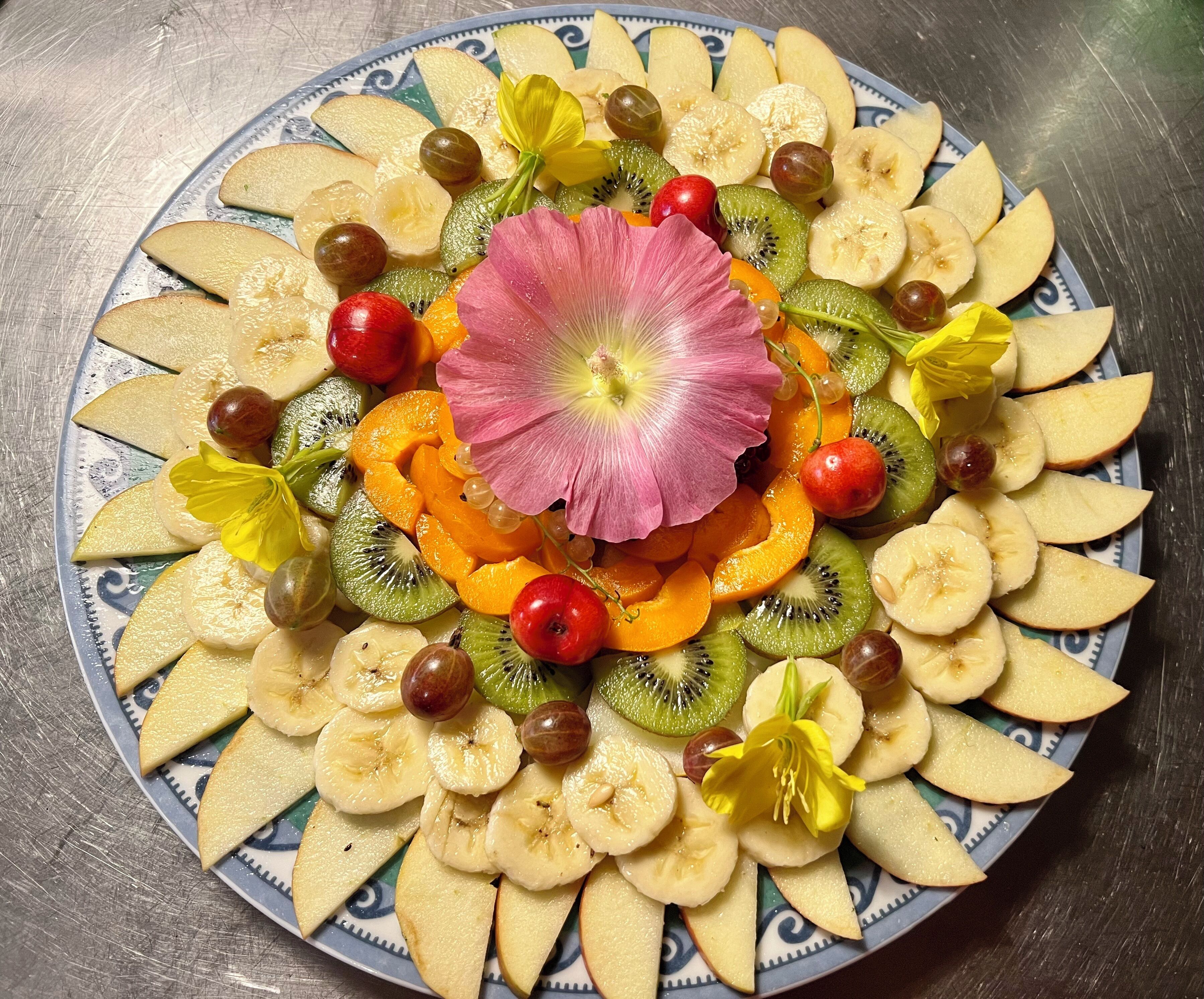 Una bandeja de frutas con rodajas de manzana, plátanos, kiwis, naranjas, uvas y una flor rosa en el centro.