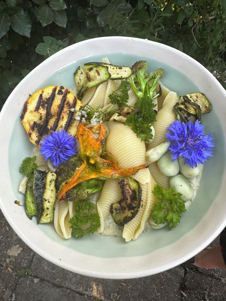 Un plato de comida con vegetales a la parrilla y pasta, adornado con perejil y flores azules.