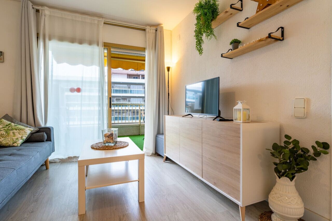 Ein modernes Wohnzimmer mit einer Couch, einem Fernseher auf einem Schrank, einem kleinen Tisch und Blick auf einen Balkon mit Pflanzen.
