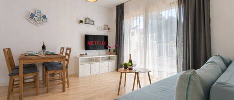 A modern living room with a wooden dining table, a sofa, a TV, and a balcony with curtains.