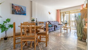 Un área de comedor con sillas de madera y una mesa redonda, un sofá con tapicería estampada, y un cuadro de una escena costera en la pared.