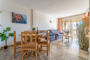 A dining area with wooden chairs and a round table, a sofa with patterned upholstery, and a painting of a coastal scene on the wall.