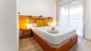 A bedroom with a wooden headboard, a bed with a white mattress and a rolled towel, a nightstand with a lamp, and a window with sheer curtains. - Salou | Modern | Cozy (5pax) Wifi | AA|Parking (Salou)