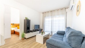 A modern living room with a grey sofa, a flat-screen TV, a white coffee table, and a small plant. - Salou | Modern | Cozy (5pax) Wifi | AA|Parking (Salou)