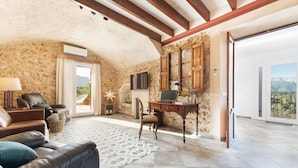 A spacious living room with a stone wall, leather sofas, a wooden desk, and a large window offering a mountain view. - Can Jaume Fuster in Buger (Buger)