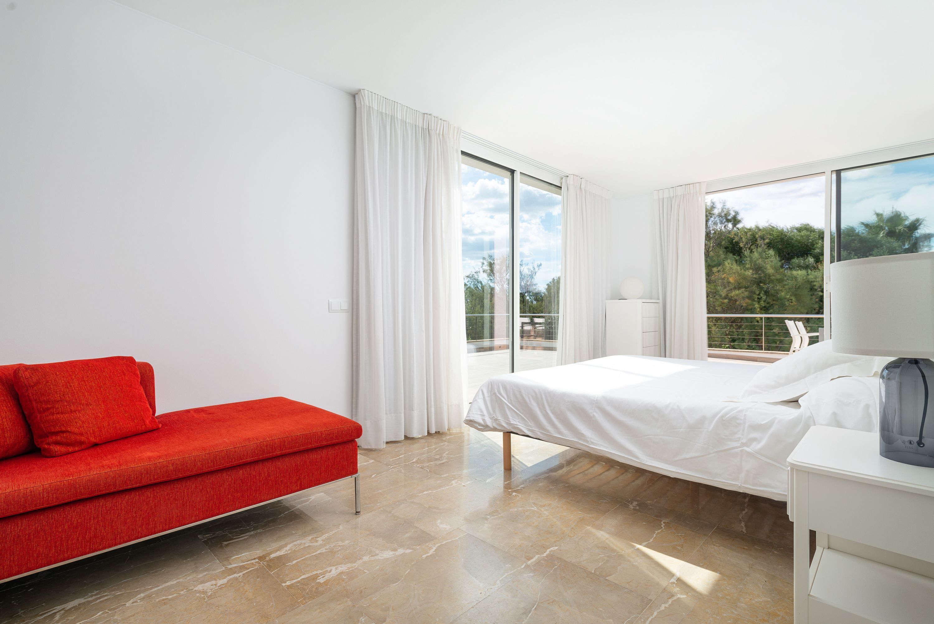 A modern hotel room with a bed, a red sofa, and large windows offering a view of the outdoors.