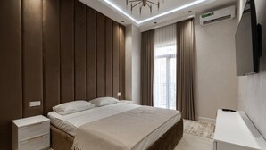 A modern bedroom with a wooden headboard, a bed with white linens, a bedside table, a TV mounted on the wall, and a window with curtains. - Tashkent city Bulevard Fontaine (Tashkent)
