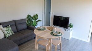 Een woonkamer met een grijze bank, een ronde tafel met twee stoelen, een flatscreen televisie en een potplant.
