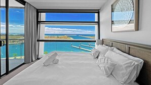 A bedroom with a large window overlooking a coastal view, a bed with white linens, and a decorative wall piece.