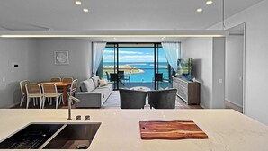 A modern living room with a large window overlooking a scenic ocean view, a dining area with a table and chairs, and a kitchen with a large island.