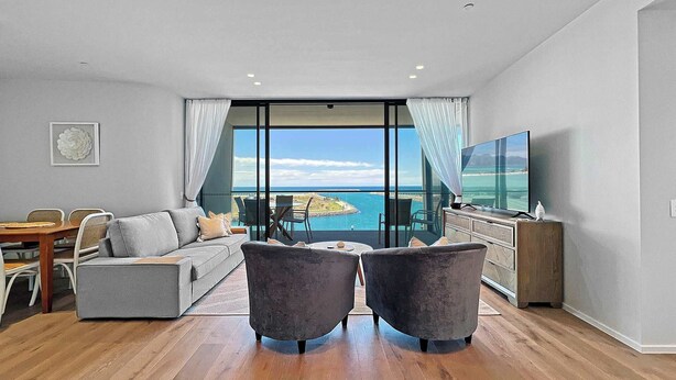 A modern living room with a sofa, two armchairs, a dining table, and a TV. It features large windows with a view of the ocean.
