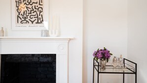A room with a white fireplace, a black brick wall, a framed abstract artwork, and a glass table with purple flowers and candles.