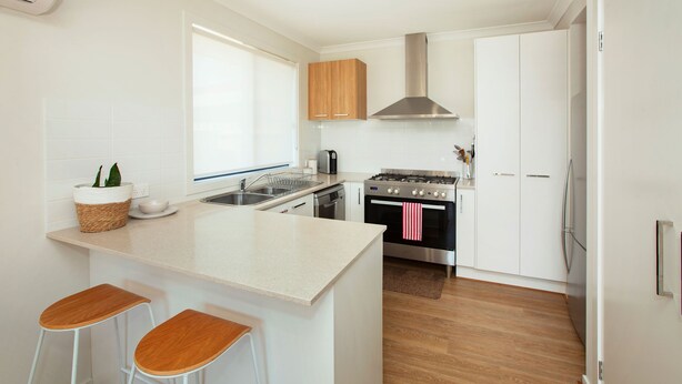 A modern kitchen with a central island, bar stools, a stainless steel stove, and wooden cabinets. - Northside Townhouse, Geelong (Norlane)