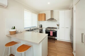 A modern kitchen with a central island, bar stools, a stainless steel stove, and wooden cabinets. - Northside Townhouse, Geelong (Norlane)